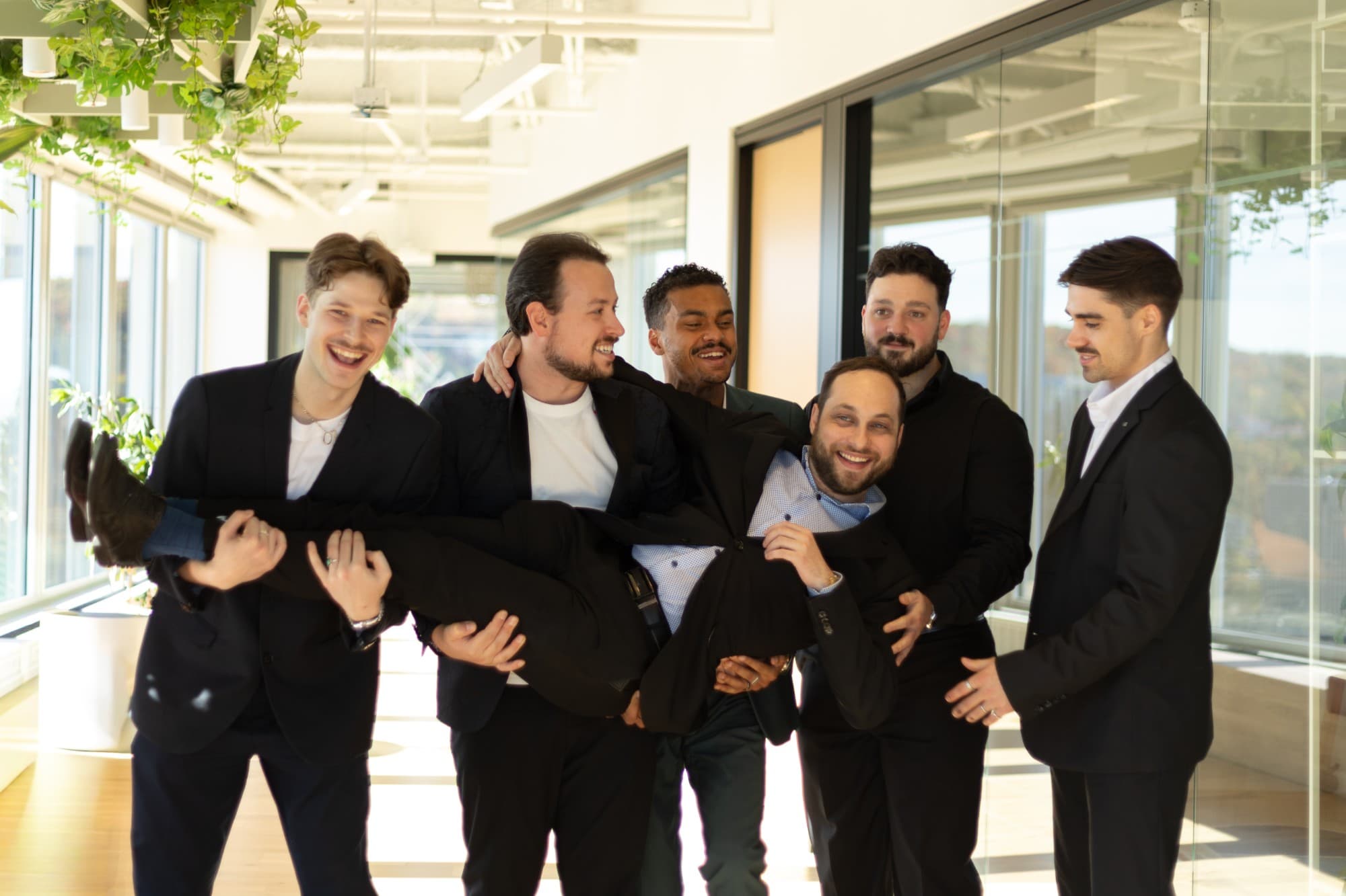 Six membres de l’équipe Omnigo.ca X Techguys, en habits sombres, rient ensemble dans un couloir lumineux en portant un collègue dans leurs bras.